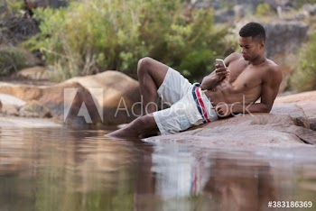 Man in swim trunks using smart phone on rocks at lakeside