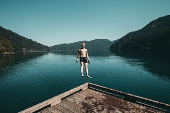 man jumping from brown wooden platform unto lake facing backwards