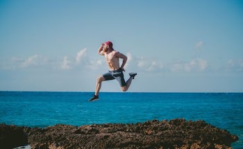 man jumping in mid air while looking ahead beside body of water during daytime