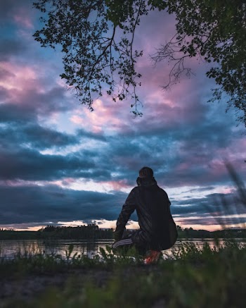 Man Kneeling and Facing Calm Body of Water Under Dramatic Clouds