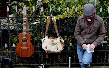 Man Leaning Against Black Steel Fence Beside White and Brown Sling Bag and Brown Acoustic Guitar
