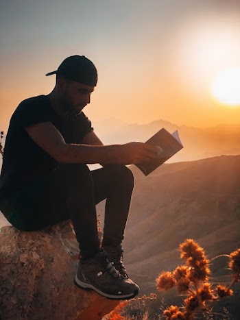 Man Sitting On A Rock Outdoors With The View Of The Mountains Reading A Book 