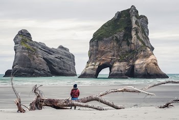 man sitting on driftwood in front of stack landform