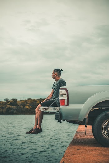Man Sitting On Vehicle Tailgate Near Body Of Water