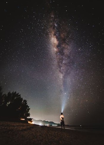 man standing on seashore wearing headlamp under milky way