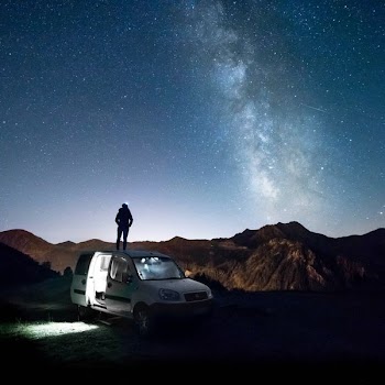 man standing on white minivan looking at mountain under milkyway