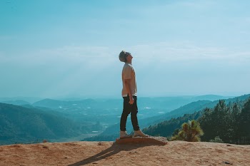 Man Wearing Beige Shirt and Black Pants Looking Up With Blue Sky and Mountains in the Background