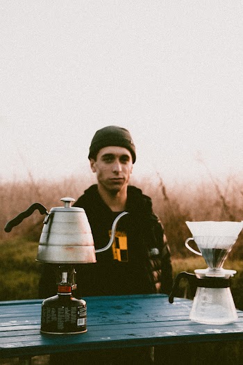 Man Wearing Black Jacket Standing While Staring on Kettle on Top of Portable Propane Tank on a Wooden Table