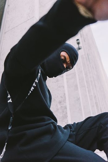 man wearing black mask, black long-sleeved shirt, and black pants sitting near wall