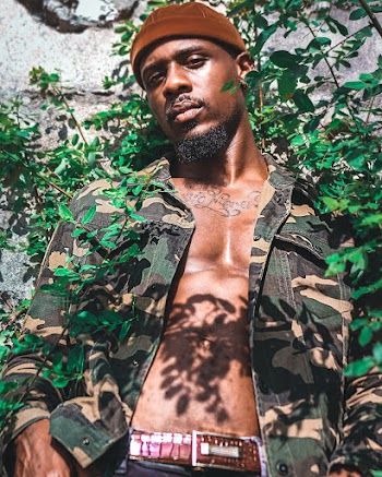 Man Wearing Brown, Green, and Black Camouflage Jacket Leaning on Gray Wall With Plants