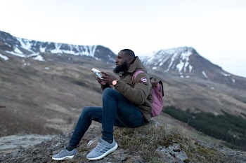 Man Wearing Brown Jacket and Blue Pants Sitting on Green Grass Holding Rectangular Case