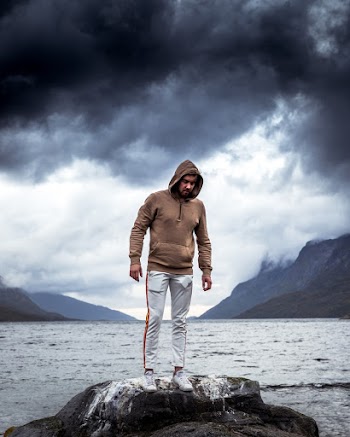 Man Wearing Brown Sweatshirt and White Sweatpants Standing on Rock Near Body of Water