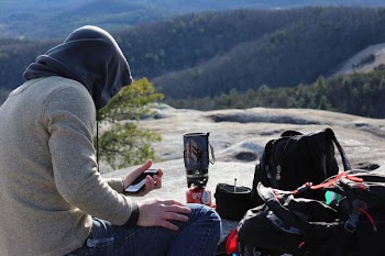 man wearing gray hooded jacket near top of mountain while using smartphone during daytime