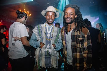 Man Wearing Hat and Tasseled Open Cardigan Standing Beside Man Wearing Plaid Button-up Shirt