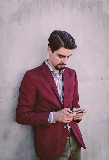 Man Wearing Maroon Blazer Leaning on Gray Concrete Wall While Using His Smartphone