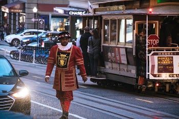 Man Wearing Red and Yellow Dress Walking on Concrete Road Near White and Red Cable Train