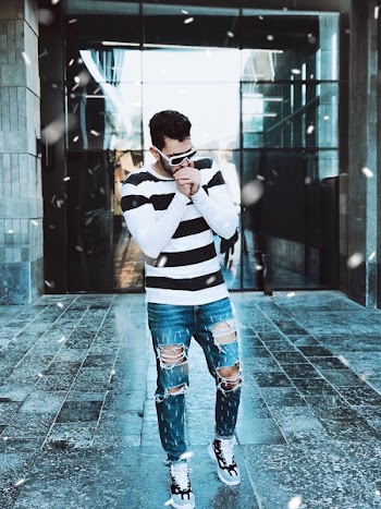 Man Wearing White and Black Striped Sweatshirt and Blue Denim Jeans Standing on Gray Surface in Front of Glass Building