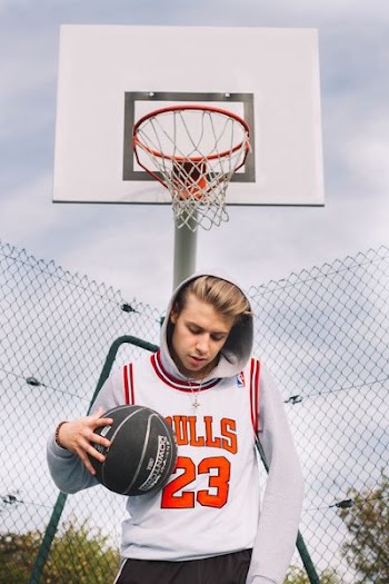 man wearing white and red Chicago Bulls Michael Jordan 23 jersey top and gray pullover hoodie while holding black basketball standing in front of white basketball system at daytime