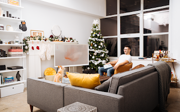 Man Wearing White Crew-neck T-shirt and Eyeglasses Holding Book While Sitting on Chair Near Christmas Tree in Living Room
