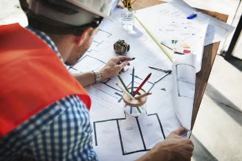 Man Wearing White Hard Hat Leaning on Table With Sketch Plans
