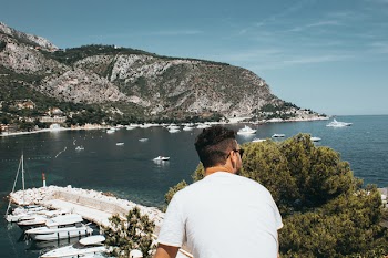 Man Wearing White Shirt and Sunglasses in Front of Boats and Mountain While Looking Right Side Under Blue Sky