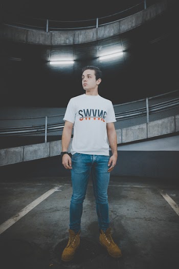 Man Wearing White T-shirt and Blue Jeans Standing on Black Concrete Floor