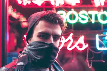man with face half covered with mask standing in front of red and green neon light signage during daytime