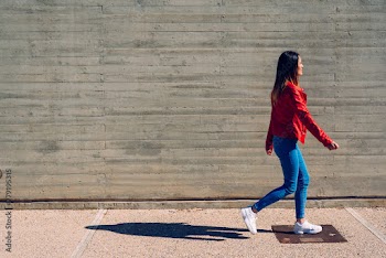 Millenial woman walking calmly in blue and red jeans next to a cement wall, gray background copy space.