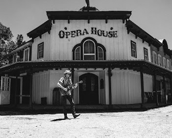 Monochrome Photo of Man Walking on Street Playing an Acoustic Guitar