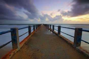 One Point Perspective Photo of Brown Concrete Bridge Leading to Body of Water With Gray Nimbus Clouds As Background during Golden Hour