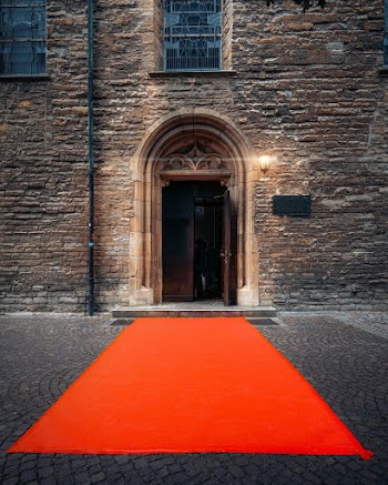 Orange Carpet Pathway Leading To The Doorway Of Brown Concrete Building With A Illuminated Wall Lamp