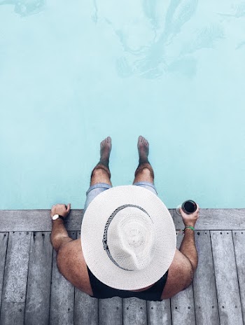 Overhead Photo of Man in White Sun Hat. Black Vest, and Denim Shorts Sitting Poolside Holding Beverage with His Feet in the Water 