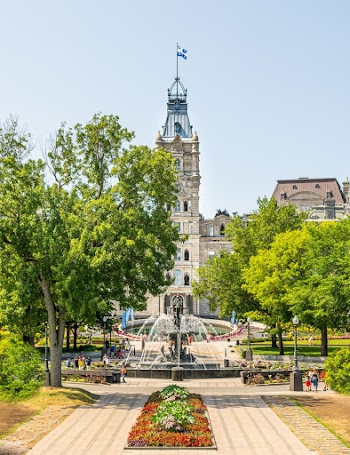 People By A Fountain In The Park With A  Walkway Surrounded With Trees And Flowers With In Front Of A Building With A Tower 
