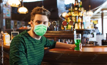 people, leisure and st patrick's day concept - young man wearing face protective medical mask for protection from virus disease drinking green beer at bar or pub