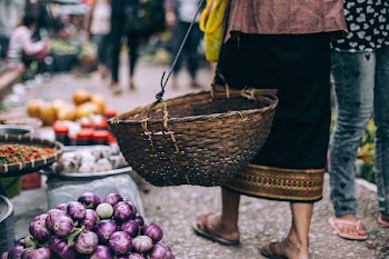 person carrying brown woven basket between person wearing gray jeans and vegetables on floor