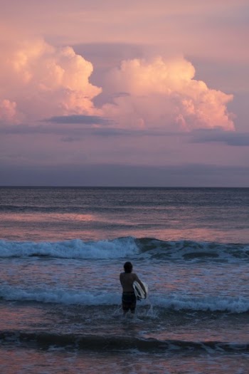 Person Carrying Surfboard Walking Against Sea Waves during Golden Hour