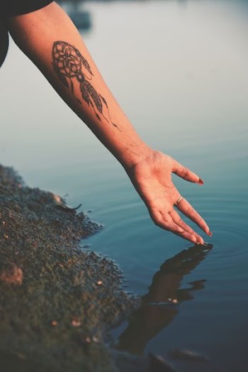 person hand reaching calm water with dreamcatcher tattoo