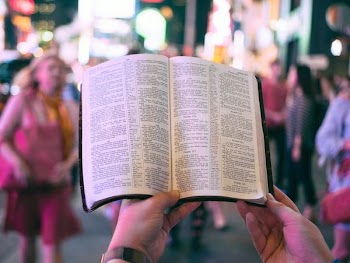 person holding bible on road with people walking on sidewalk beside buildings during nighttime