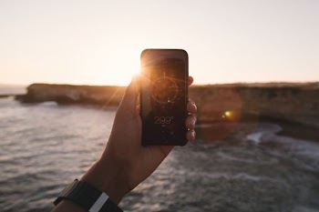person holding black phone beside body of water under white sky during golden hour