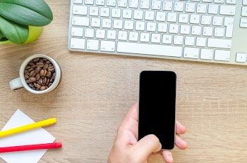 Person Holding Black Phone Near Apple Magic Keyboard on Brown Surface