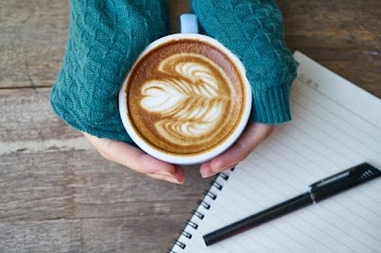 Person Holding Cup of Coffee Latte Near Notebook and Pen