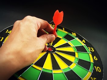 Person Holding Red Dart on Green Yellow and Black Dart Board