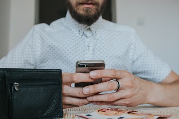 Person Holding Silver Iphone Beside Table With Black Leather Wallet and Two 10 Banknotes