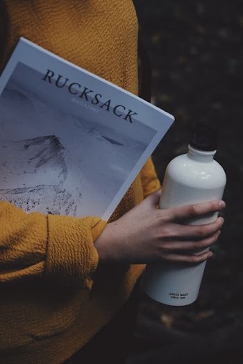 person holding white tumbler and a Rucksack magazine in between arm