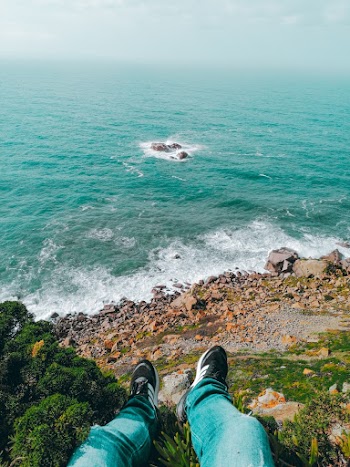 Person in Black Pants and Black Hiking Shoes Sitting on Rocky Shore