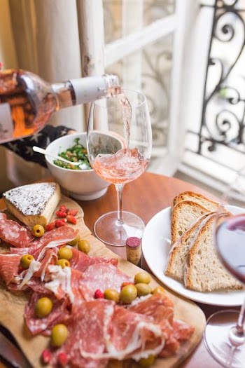 person pouring beverage on clear long-stem wine glass beside white ceramic plate with sliced bread and brown wooden board with bacons