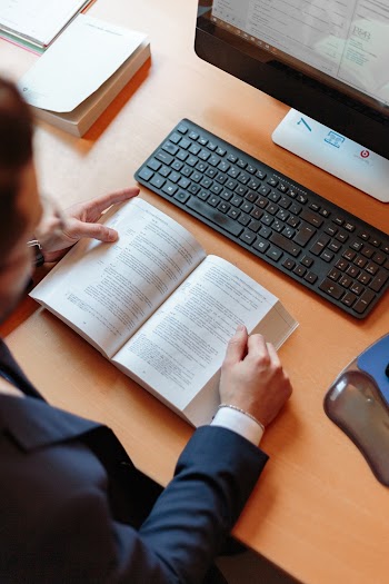 Person Reading Book With Desktop Computer Set on Table