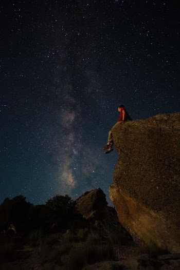 person sitting on edge of large rock under starry night sky