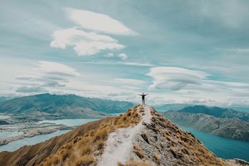person standing on peak of the mountain near lake under white cloud blue skies
