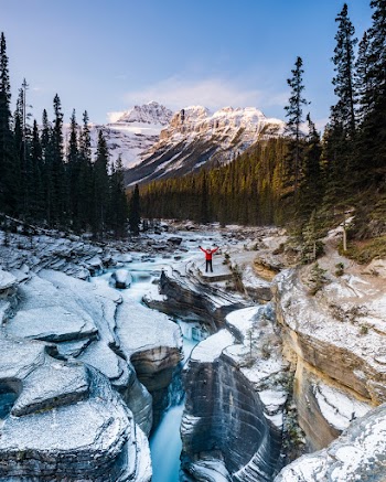 Person Standing on River Across Snow Capped Mountains